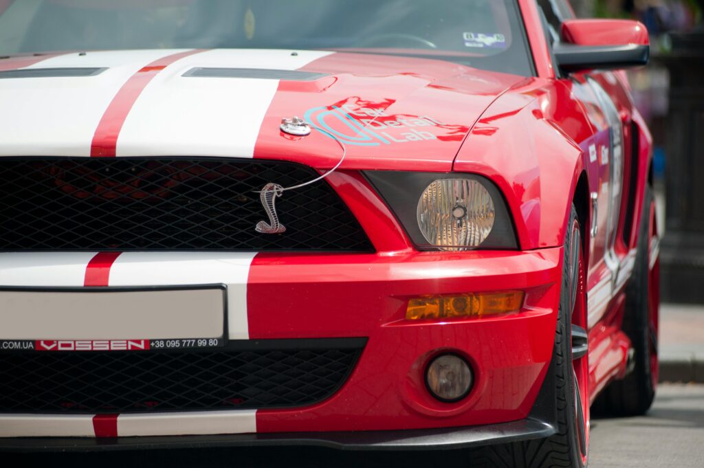 Front view of a red Ford Mustang sports car with white racing stripes in a city setting.